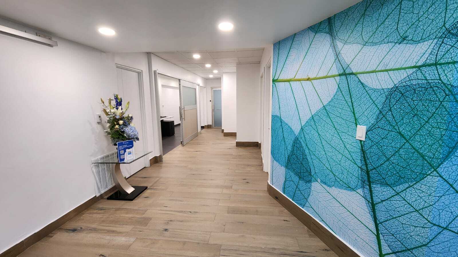 Pasillo de hospital con pared decorativa de hojas azules, mostrador con flores y puertas de tratamiento